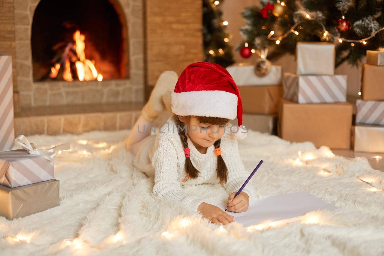 Little cute female child lying on floor on carpet and writing letter to Santa Claus, looking at empty paper and think, posing against present boxes, xmas tree and fire places.