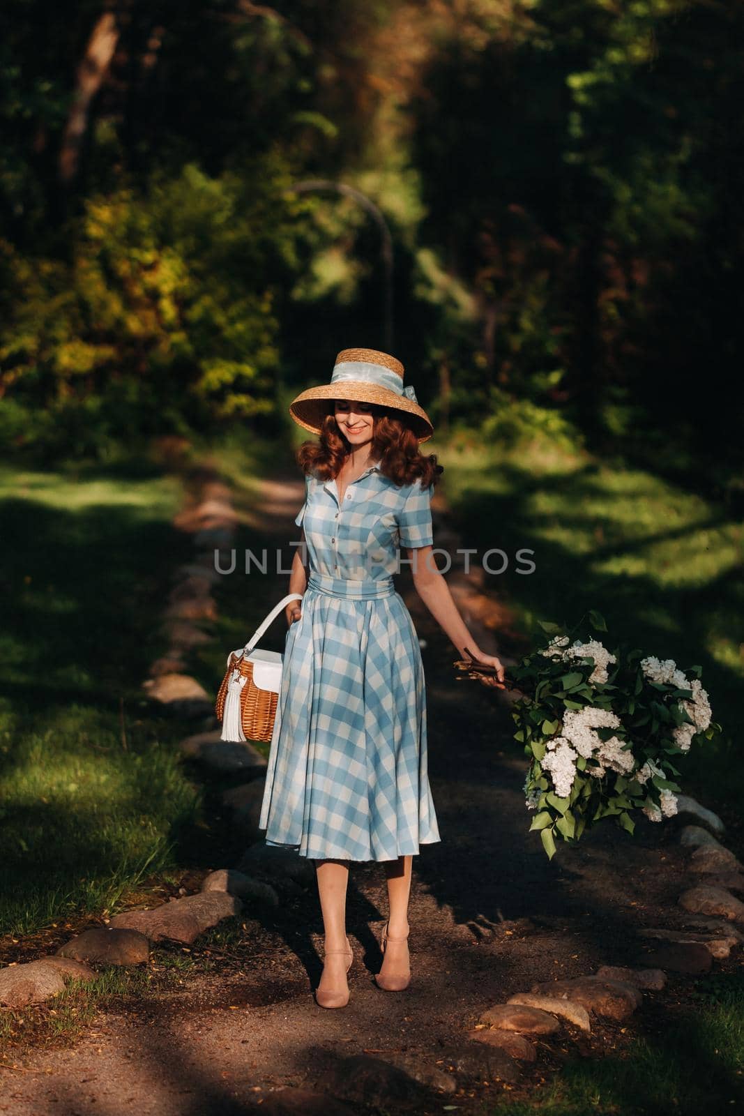 Portrait of a beautiful girl with long hair, a straw hat and a long summer dress with lilac flowers in the garden.