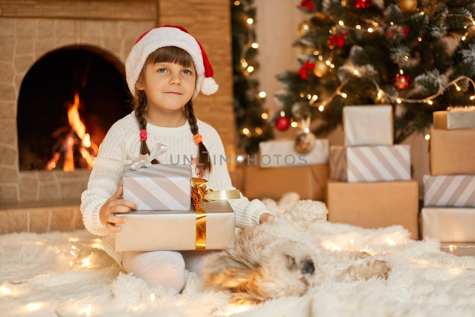 Pensive little child sitting on floor and looking away, holding gift boxes with ribbon, imaging her present, posing in decorating living room with garland, christmas tree and fireplace.