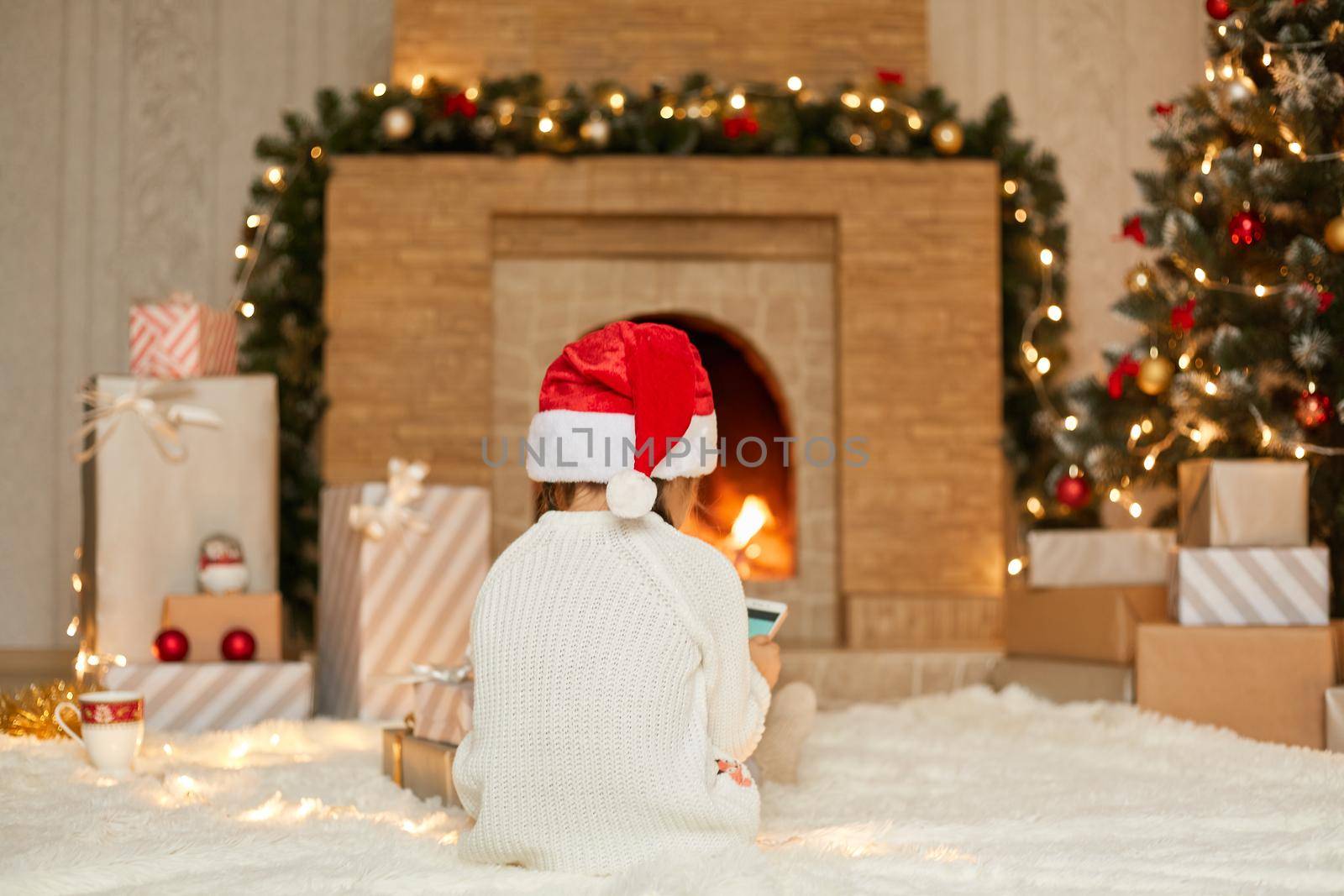 Little girl wearing santa claus hat and white jumper communicates with relatives on phone, posing backwards while sitting on floor on carpet with phone in hands. Christmas eve.