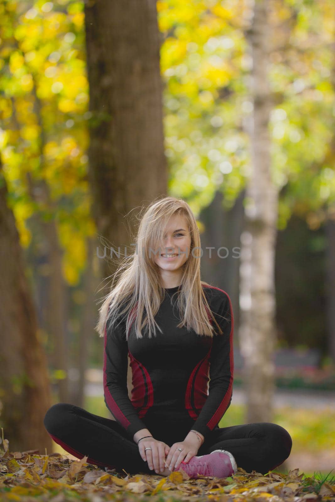 Young attractive fitness female sitting cross-legged at workout in autumn park, smiling and posing by Studia72