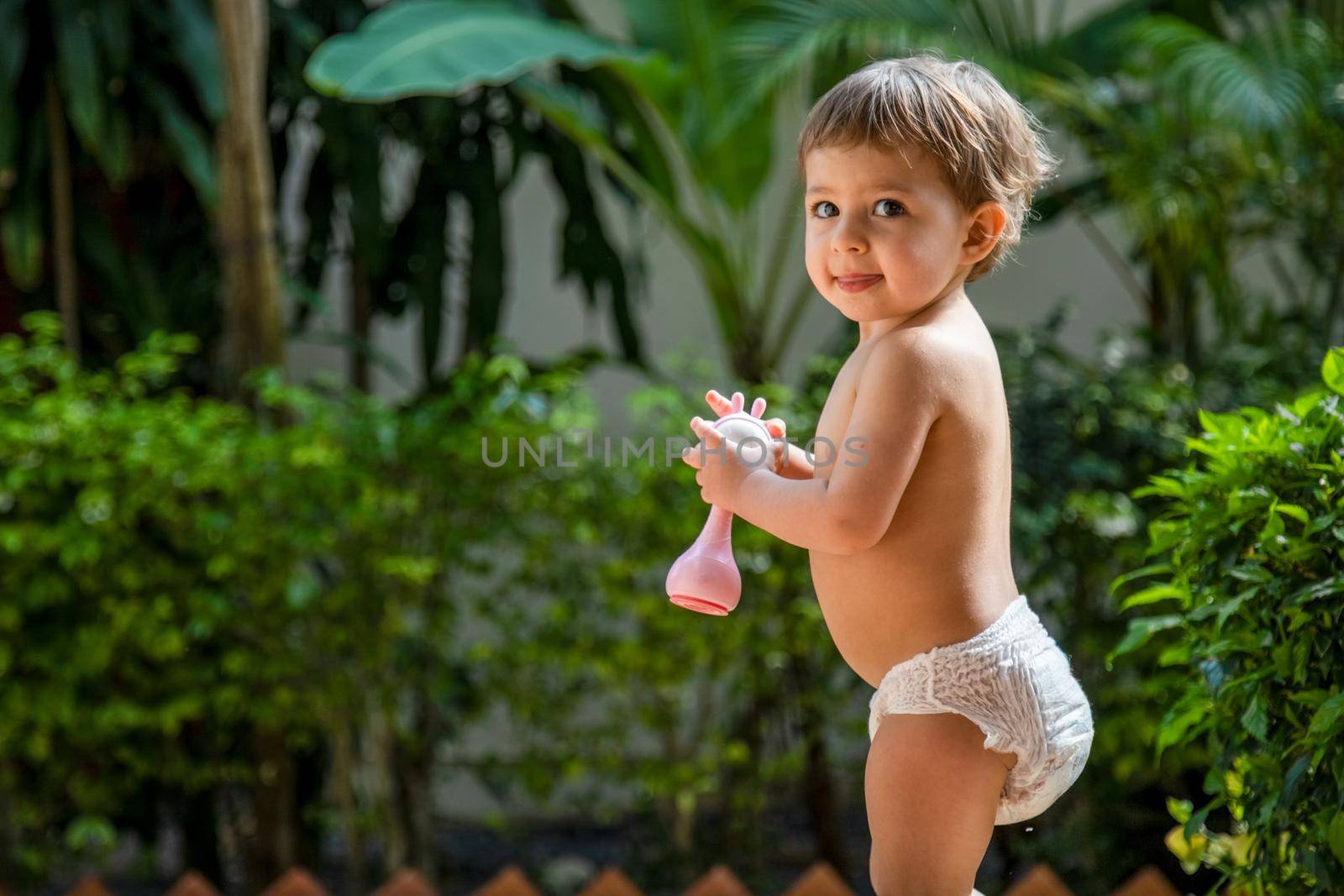 adorable toddler stands with a toy in his hands against a background of greenery in sunshine