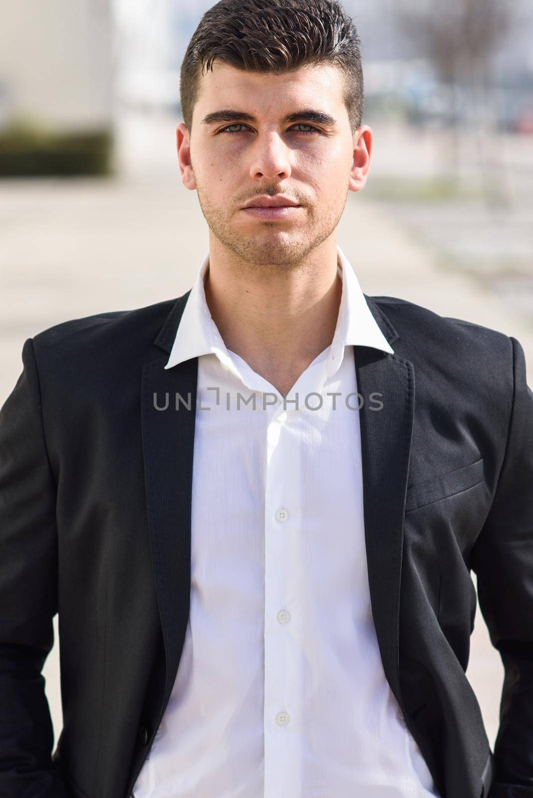 Young businessman near a modern office building wearing black suit and white shirt. Man with blue eyes