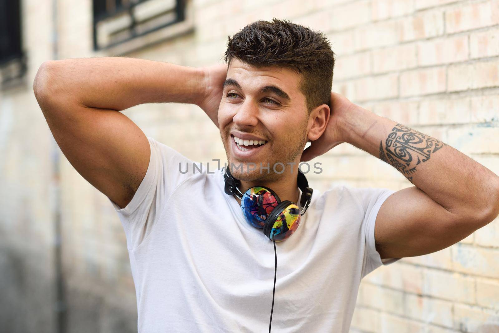 Portrait of young man in urban background smiling with headphones. Wearing white t-shirt near a brick wall