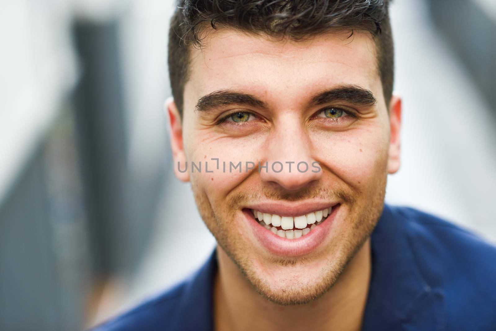 Good looking young man with blue eyes smiling in the street. Model of fashion in urban background wearing white t-shir and blue jacket
