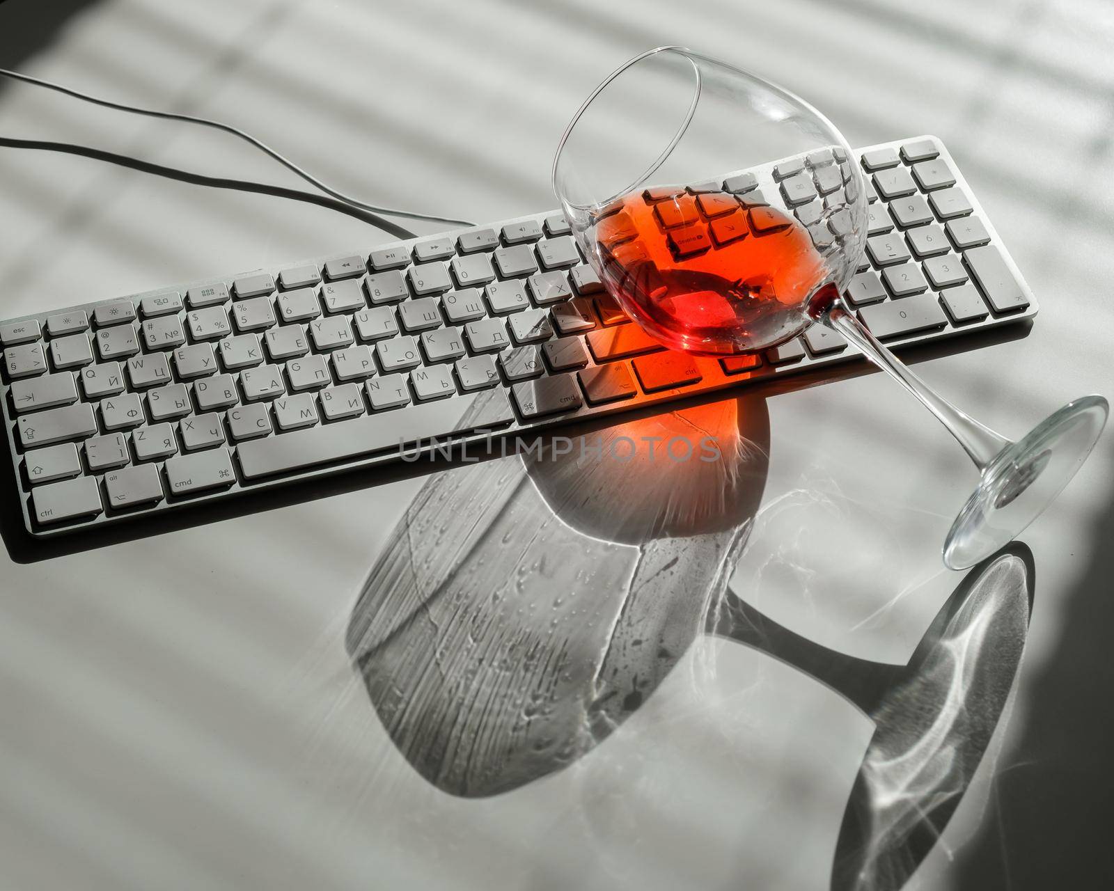 A glass of red wine lies on the keyboard on a white table with a shade from the blinds by mrwed54