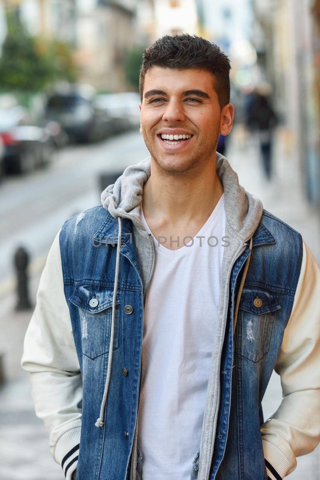 Young man with blue eyes and white teeth in the street. Model of fashion smiling in urban background wearing white t-shirt, jeans and blue jacket