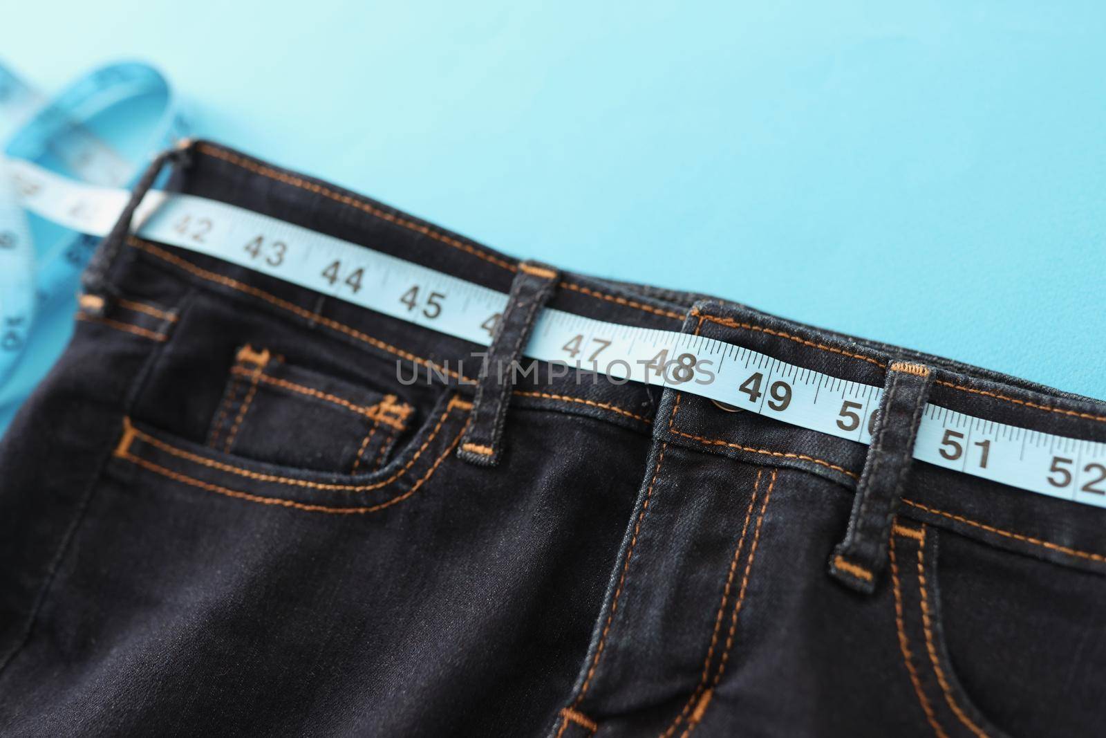 Close-up of upper part of denim trousers, jeans with blue measure tape instead of belt. Individual clothes sew in atelier. Healthy lifestyle, dieting, weight loss, tailoring concept