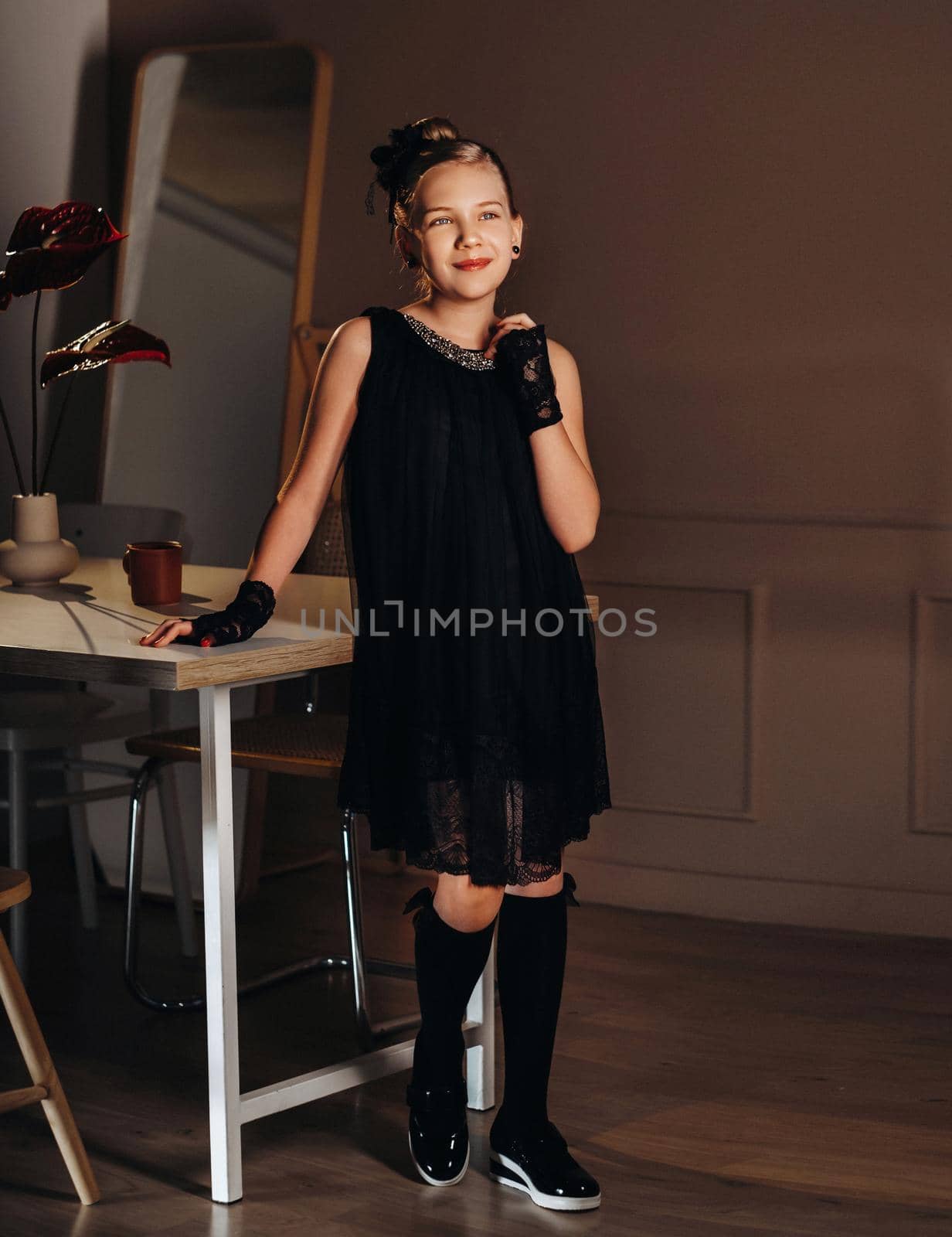 A stylish little girl in a black dress stands in the interior near the table.