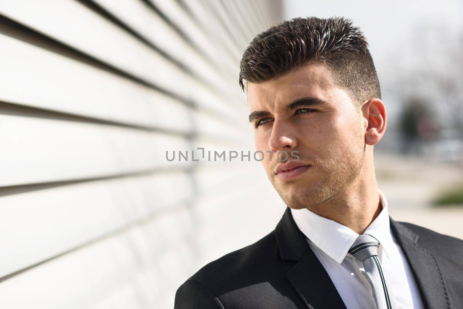 Young businessman near a modern office building wearing black suit and tie. Man with blue eyes