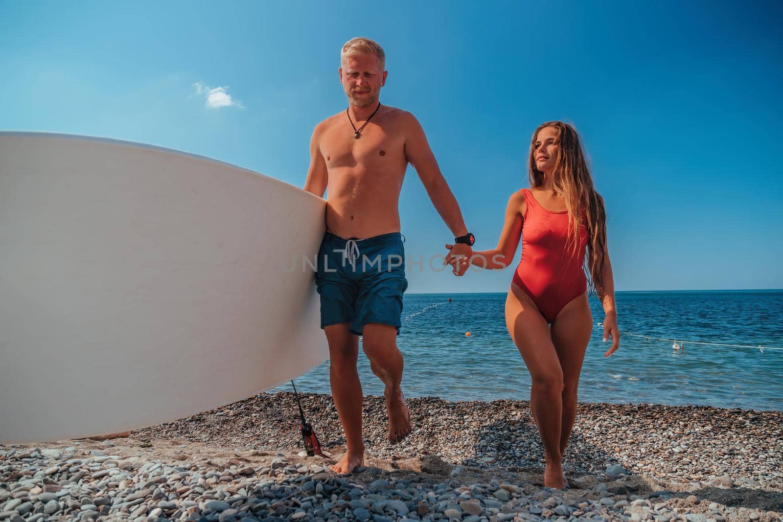 Vacation couple walking on beach together in love holding hands. Happy young couple: man in shorts holding sup board and girl in a red bikini. Beauty world, nature and outdoors travel concept.