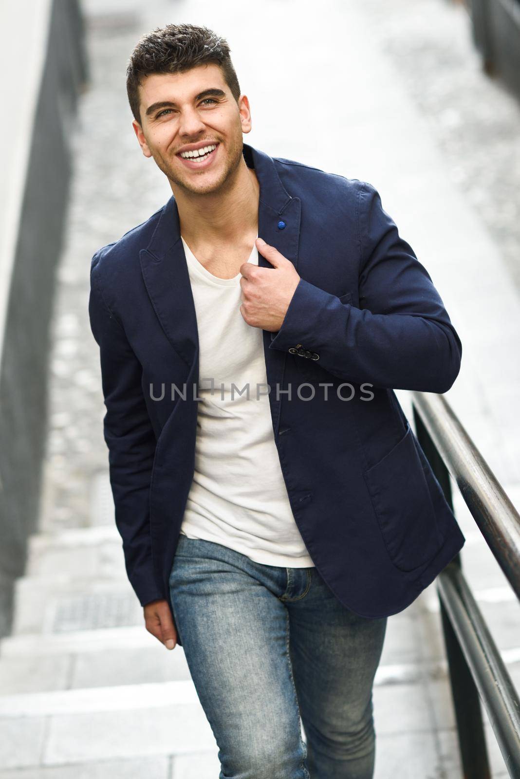 Handsome young man smiling with blue eyes in the street. Model of fashion walking in urban background wearing white t-shirt, jeans and blue jacket