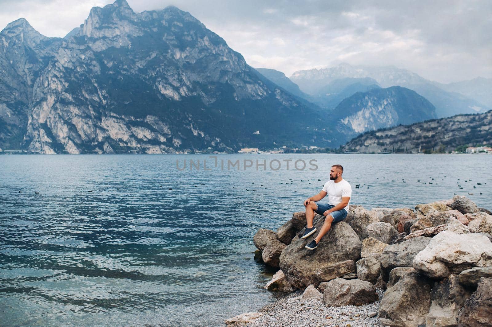male traveler sitting on the rocks against the backdrop of the Alps and lake Garda. holiday in Italy by Lobachad