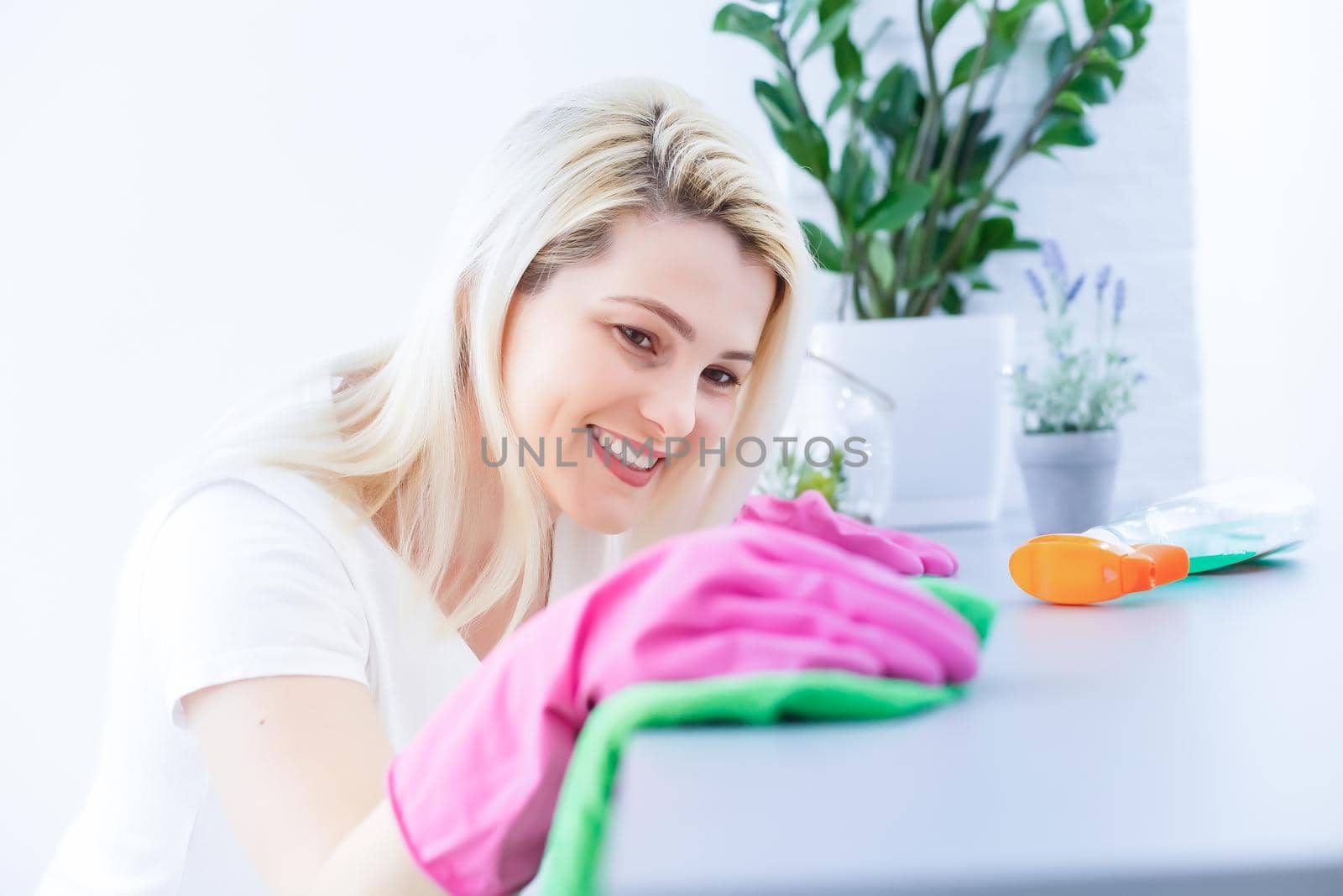 Woman in protective gloves is smiling and wiping dust using a spray and a duster while cleaning her house, close-up by Andelov13