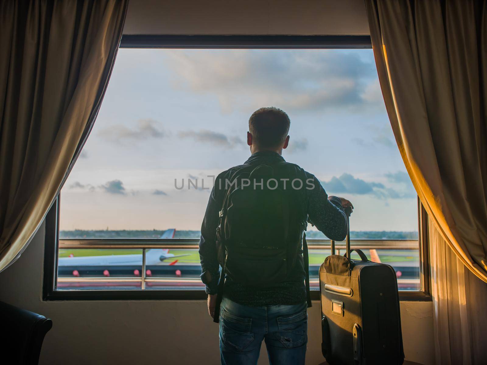 A young man watches the airplane take off from the window of his hotel room. by DovidPro