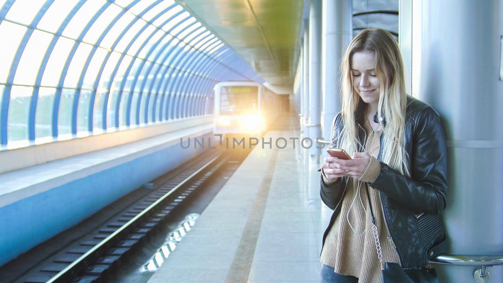 Young attractive girl with gadget long blonde hair in leather jacket with straightens hair standing in metro against the background of a train coming, perspective, horizontal