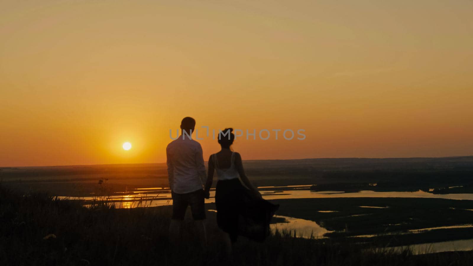 loving couple - brave young man and beautiful girl stands on high hill and looking to sunset silhouette, slider shot, wide angle