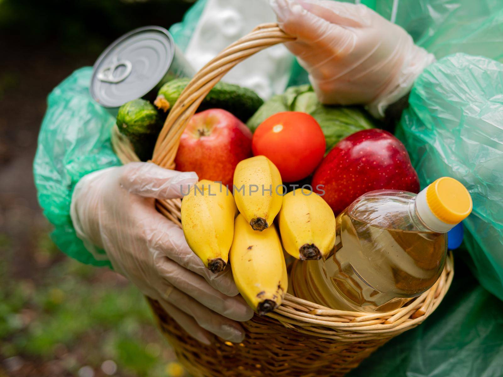 Volunteer food delivery in any weather concept. Portrait of a basket of food in the hands of a woman volunteer in protective gloves and rain coat, close-up.