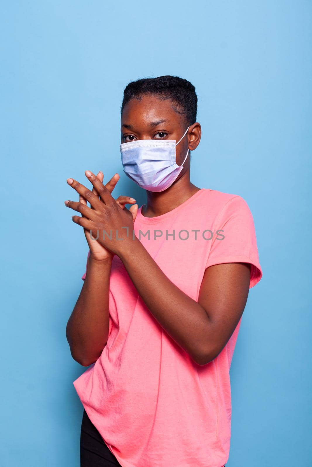 Portrait african american teenager with protective medical face mask to prevent infection with coronavirus clapping and applauding happy and joyful in studio with blue background. Celebration concept