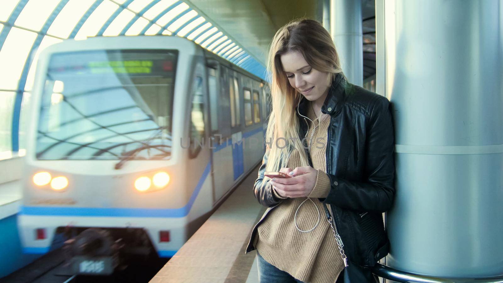 Attractive girl with gadget long blonde hair in leather jacket with straightens hair standing in metro against the background of a train coming, horizontal