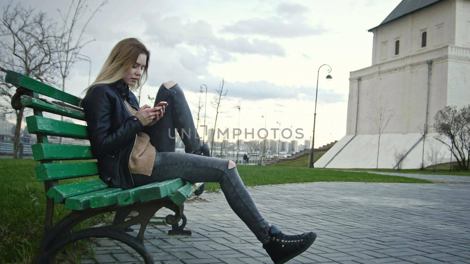 Cute happy girl with long blonde hair in leather jacket straightens hair use gadget sitting on the bench, horizontal