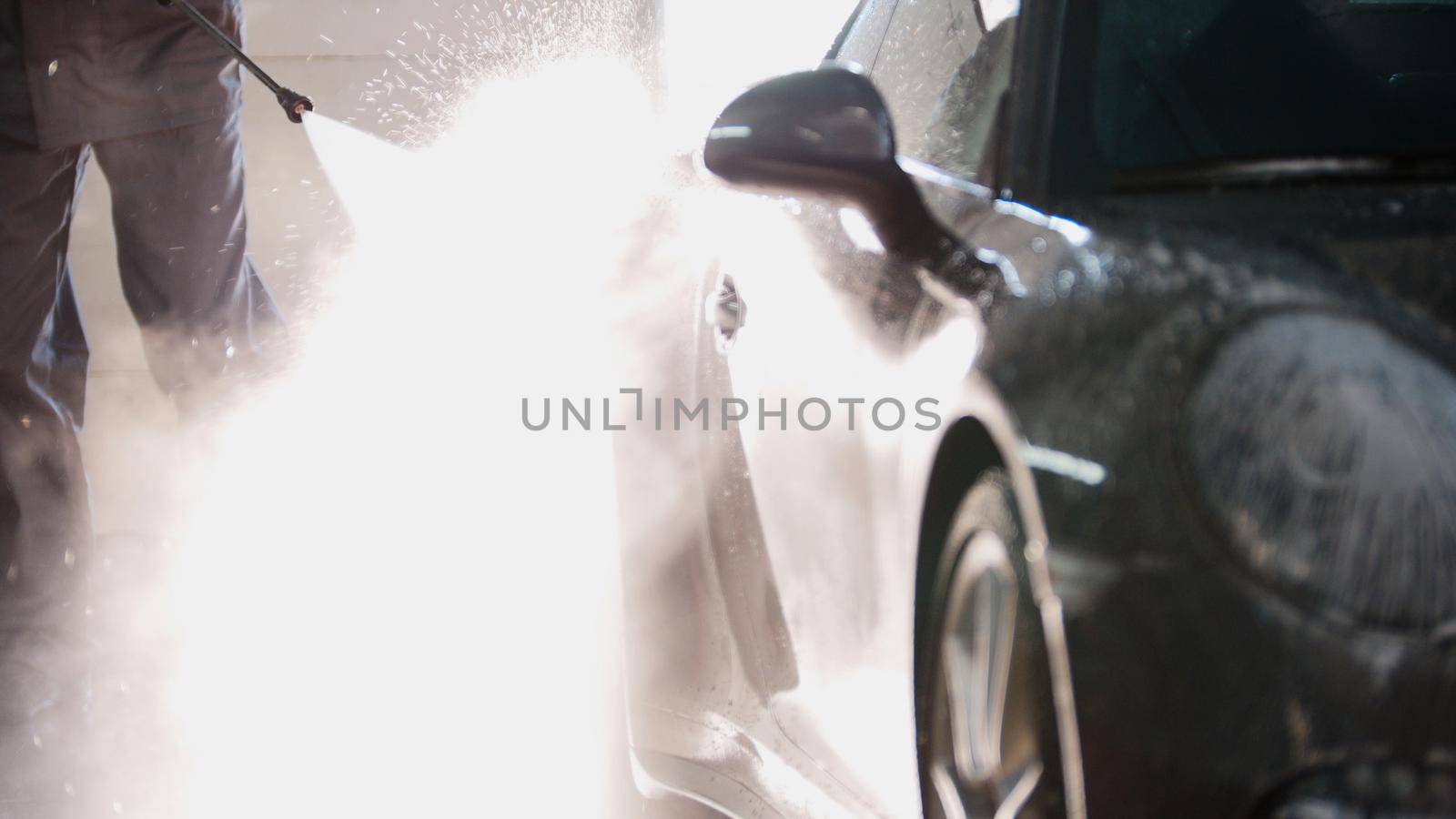Mechanic in car service is washing sport car in the suds by water hoses, telephoto, backlight