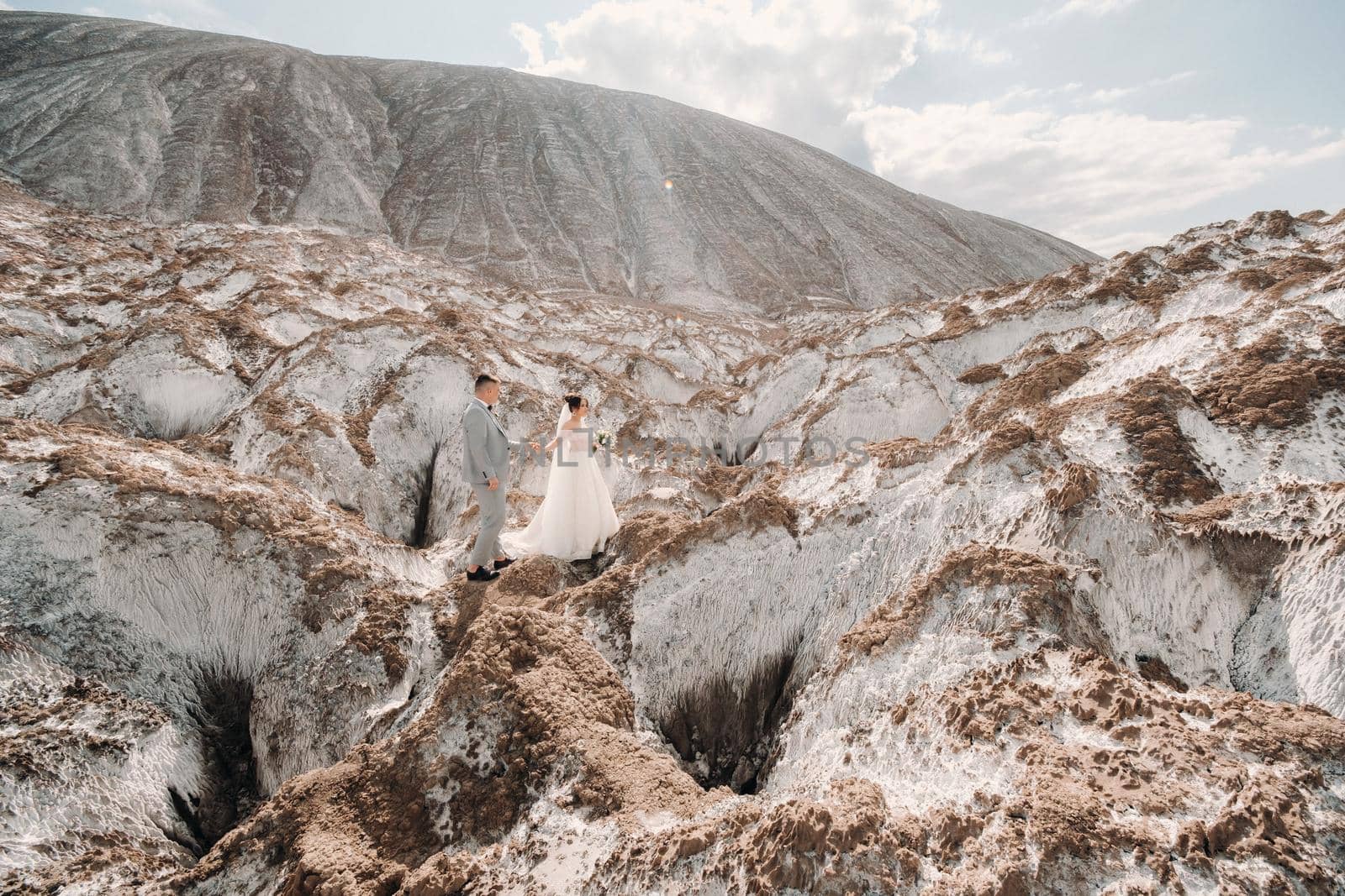 A beautiful couple of lovers posing in a white salt mountain. A young woman in a stylish wedding dress and a beautiful stylish man in a gray suit. The concept of the wedding day. by Lobachad