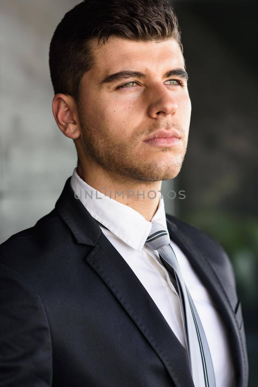 Young businessman near a modern office building wearing black suit and tie. Man with blue eyes