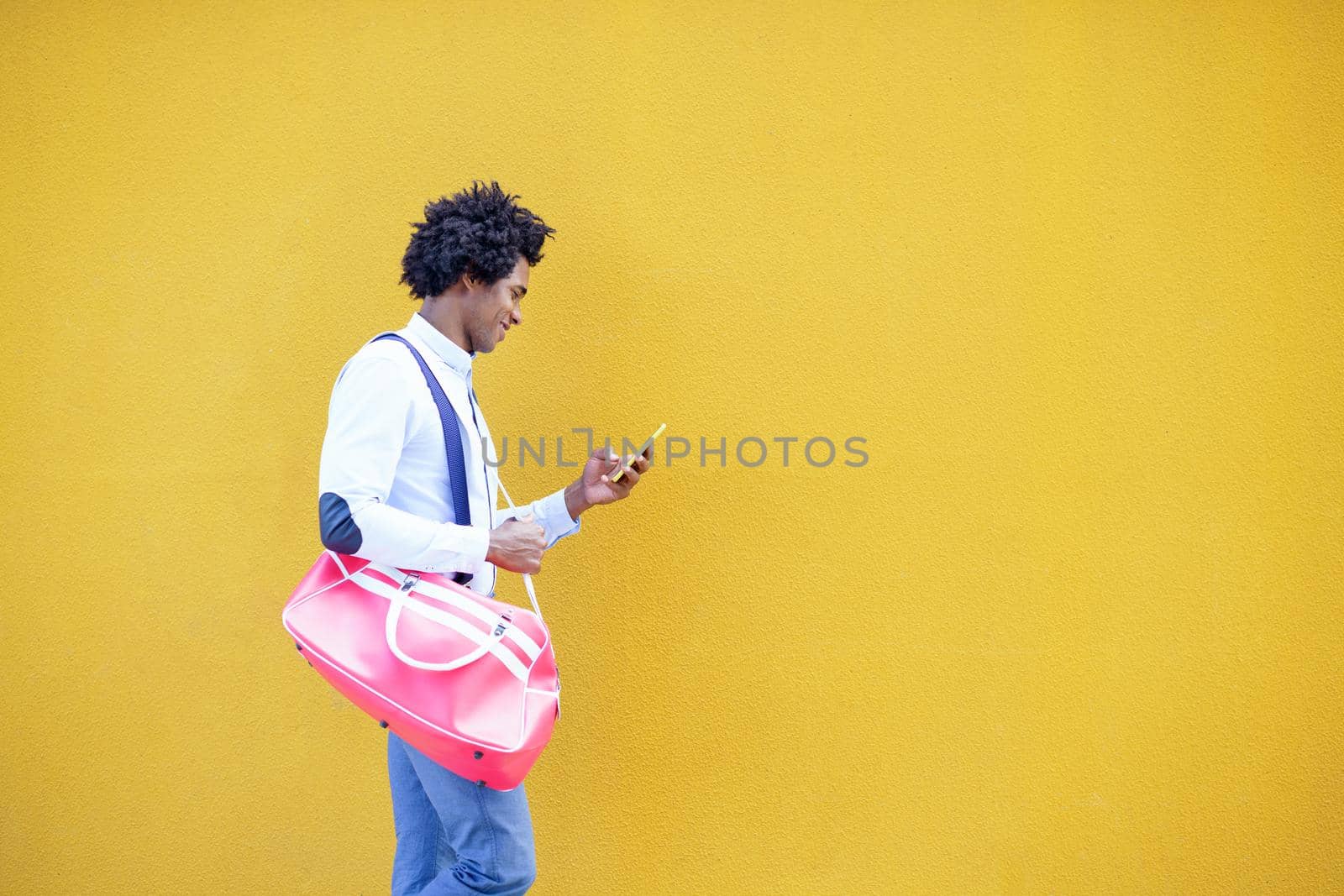 Black man with afro hairstyle carrying a sports bag and smartphone against a yellow urban background. Guy with curly hair wearing shirt and suspenders.