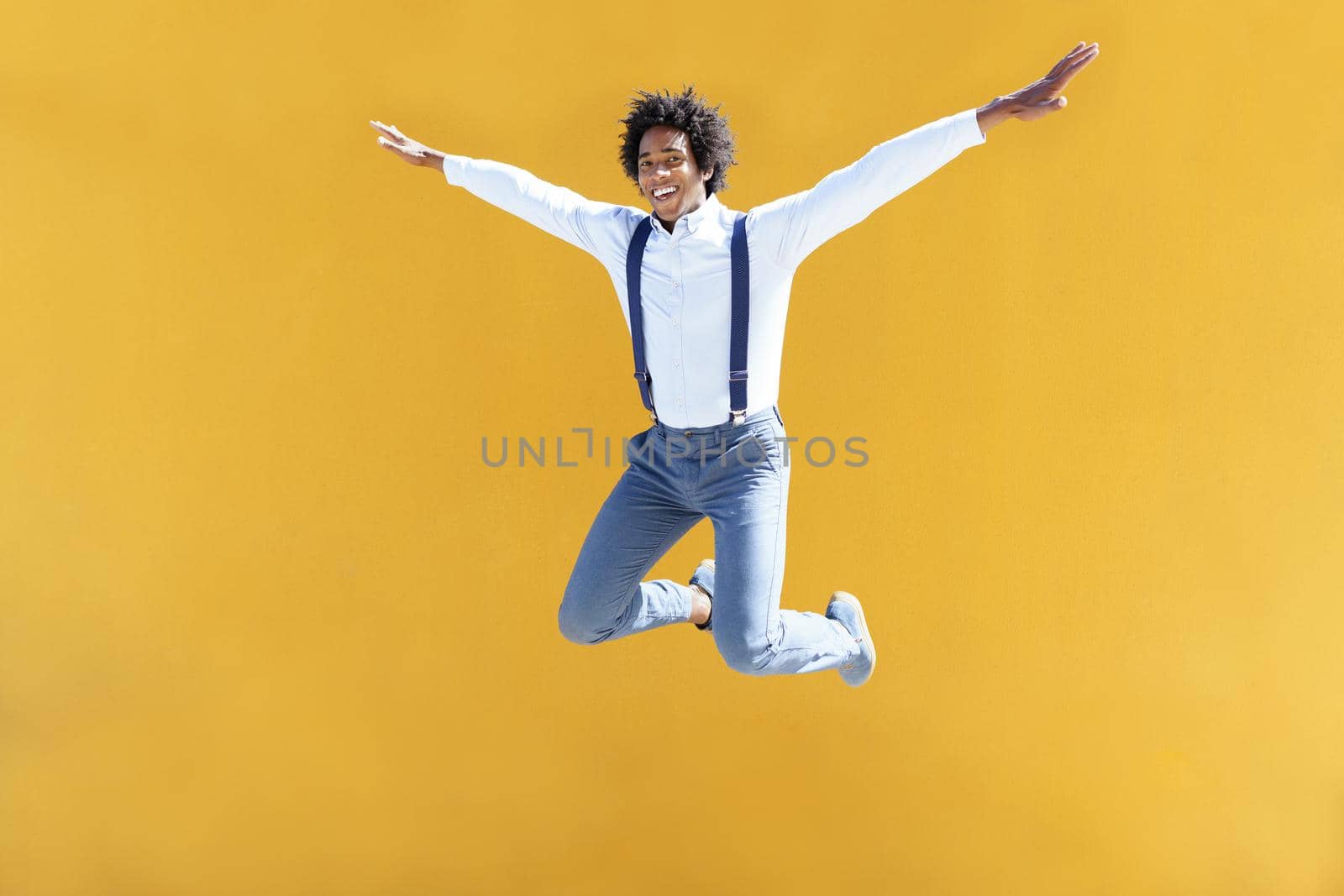 Black man with afro hair jumping on a yellow urban background. Guy wearing shirt and suspenders.