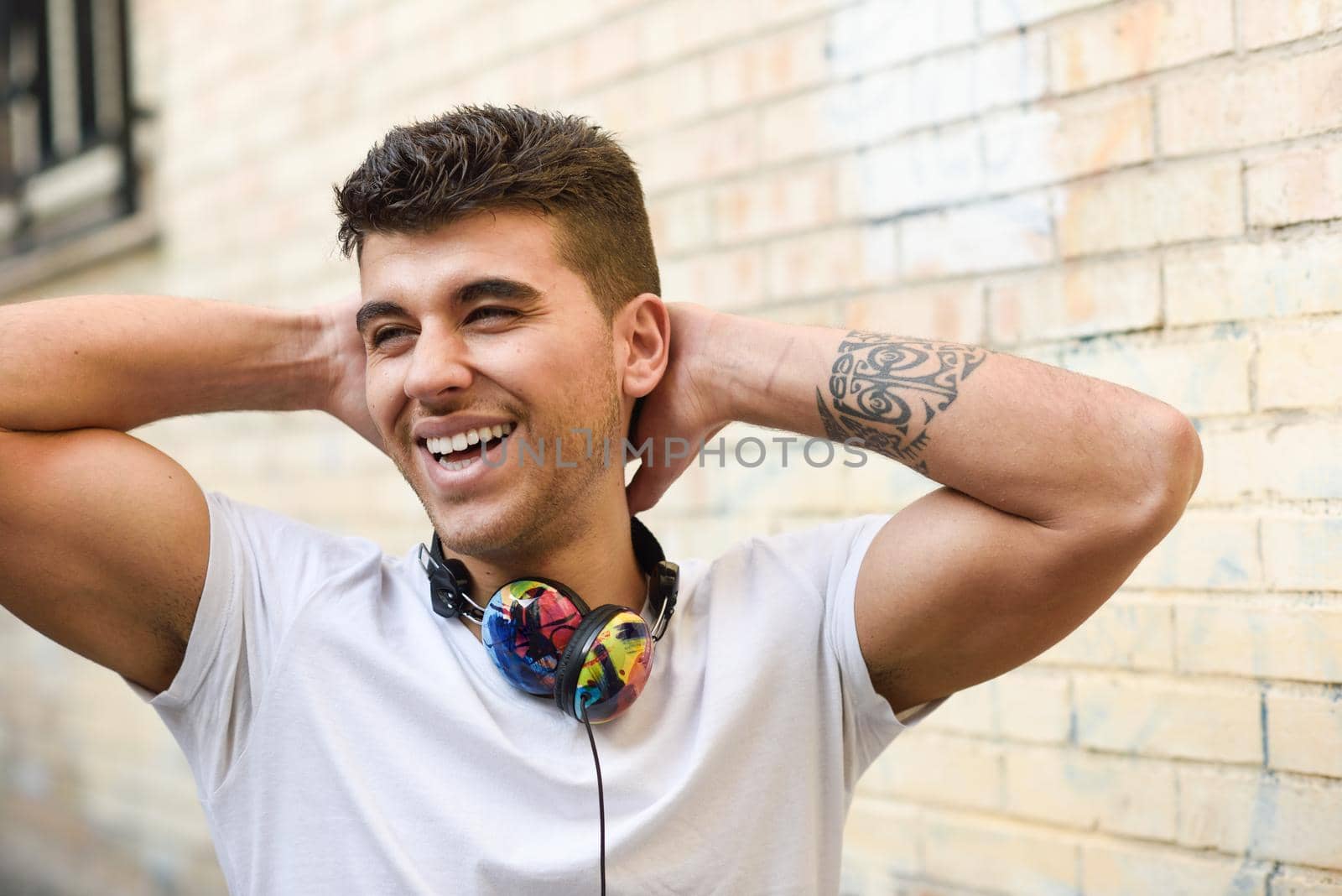 Portrait of young man in urban background smiling with headphones. Wearing white t-shirt near a brick wall