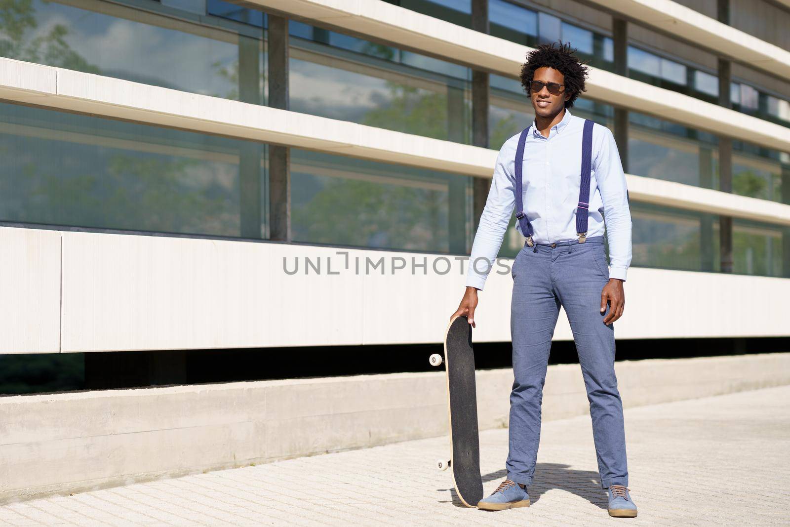 Black male worker with afro hairstyle standing next to an office building with a skateboard and sunglasses.