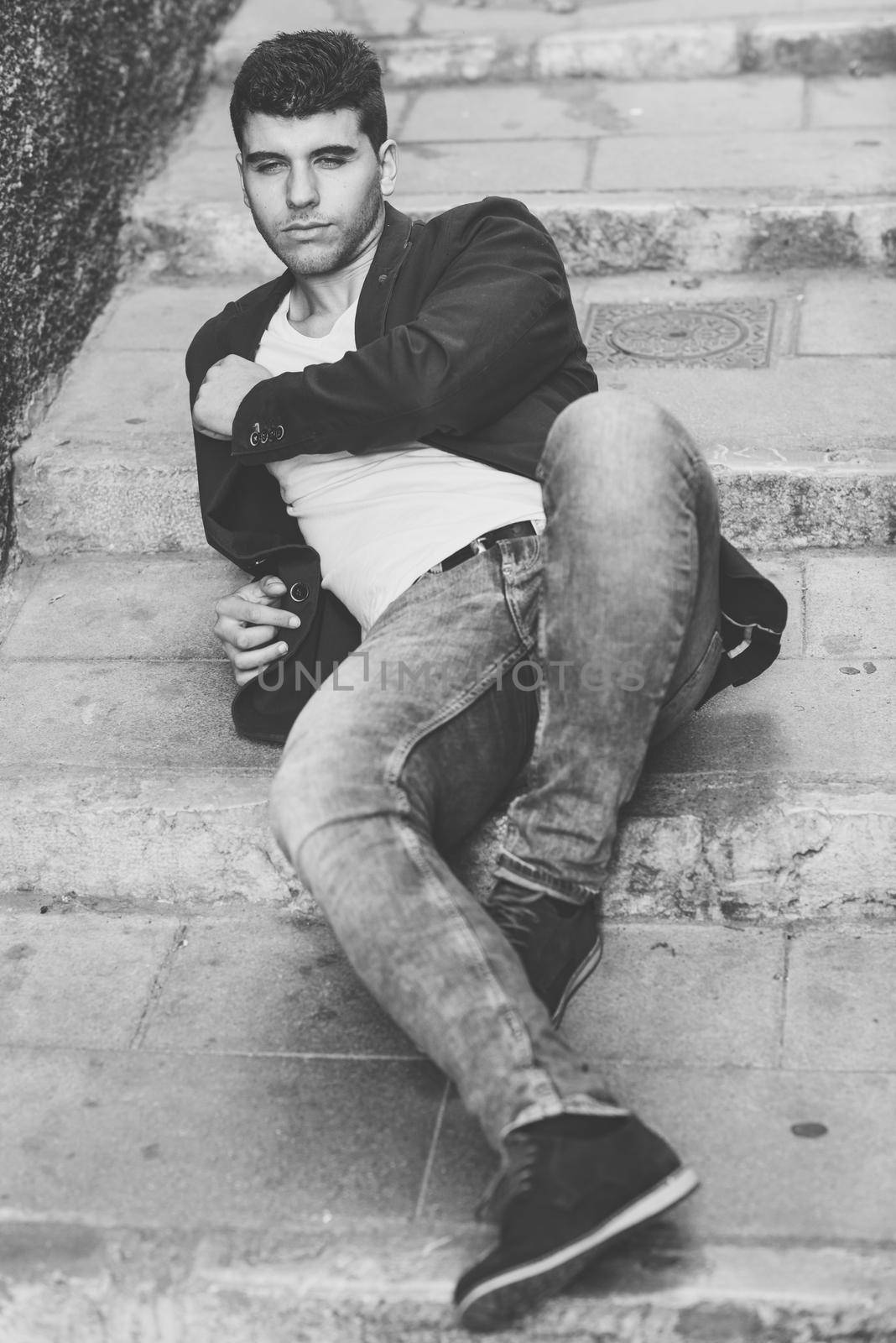 Good looking young man with blue eyes in the street. Model of fashion sitting in urban stairs wearing white t-shirt, jeans and blue jacket