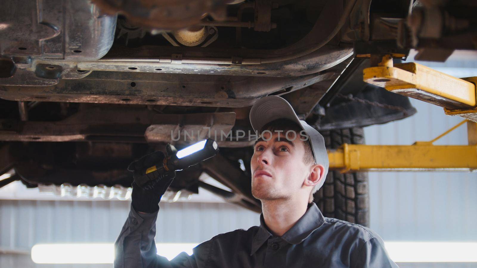 Mechanic with thr lamp is checking the bottom of car in garage automobile service, close up, telephoto