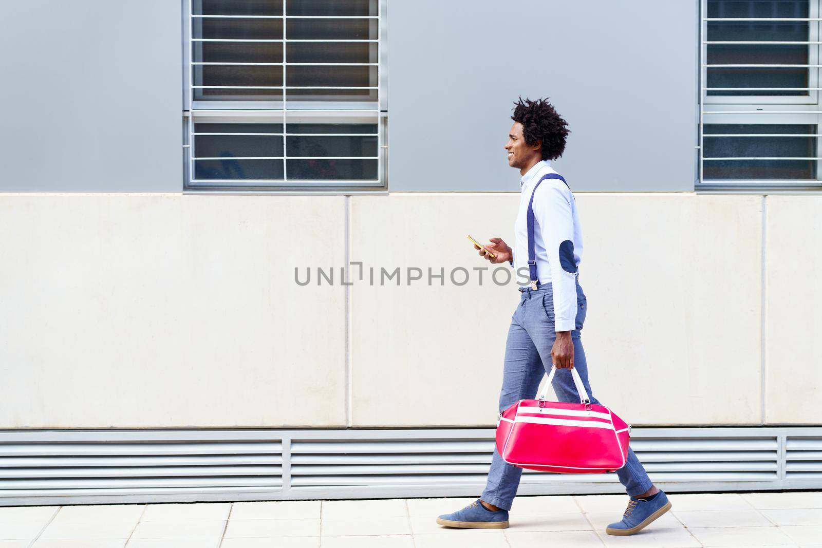 Black man with afro hairstyle carrying a sports bag and smartphone in urban background. Guy with curly hair wearing shirt and suspenders.