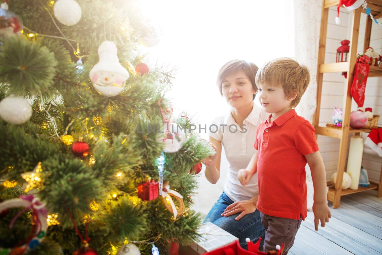 Smiling mother with her beautiful little son decroating Christmas tree with baubles at home.