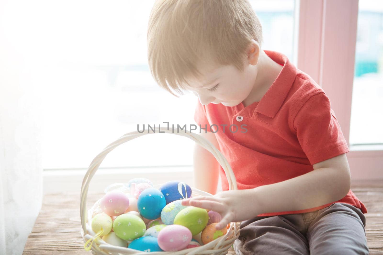 Little cute boy sits by the window with a full basket of colorful Easter eggs. .