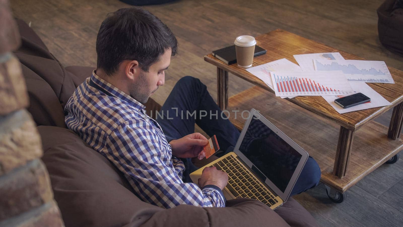 A man in a plaid shirt sitting in the seat bag and makes a purchase on the Internet. In the hands of a credit card and a laptop computer.