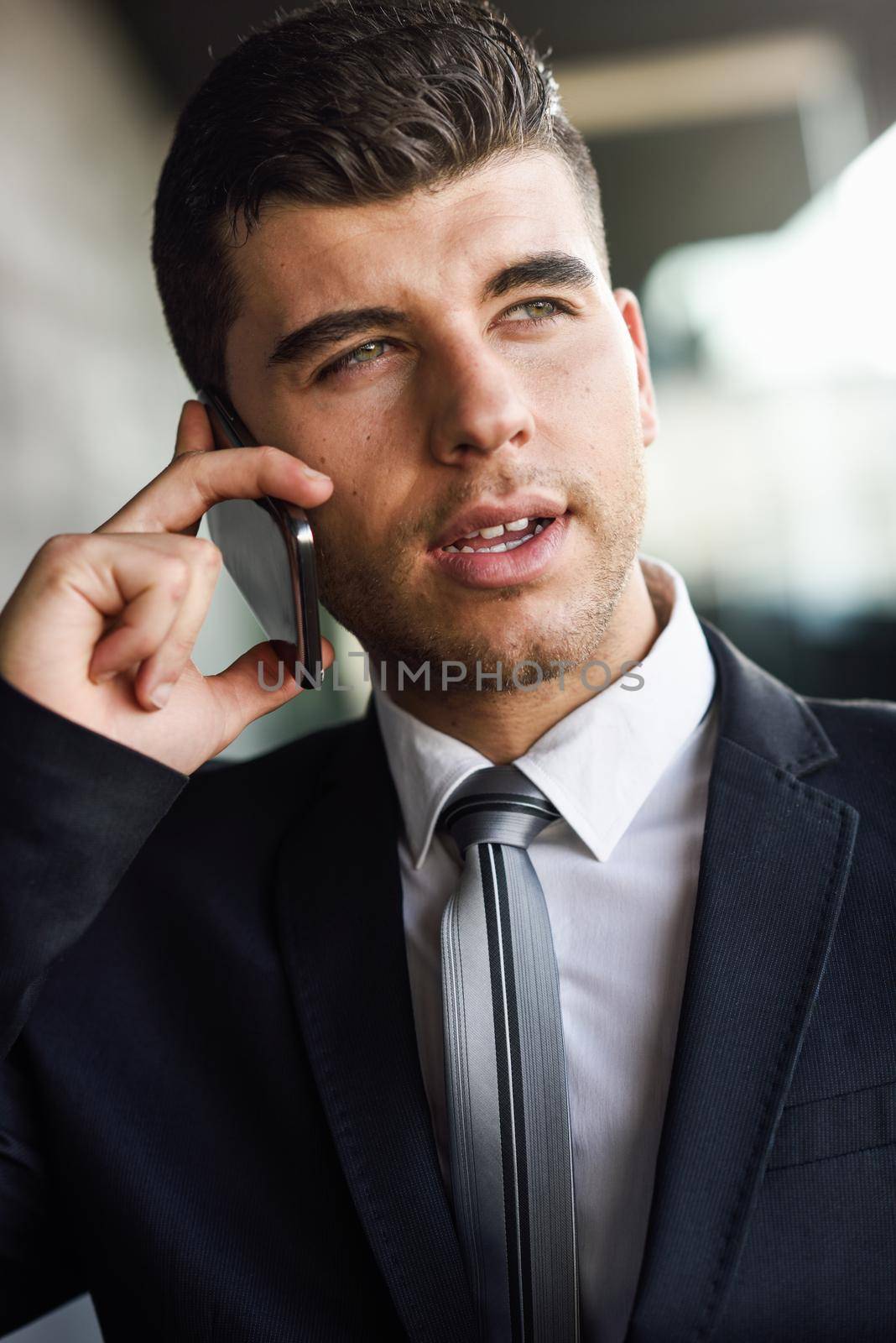 Attractive young businessman on the phone in an office building wearing black suit and tie. Man with blue eyes