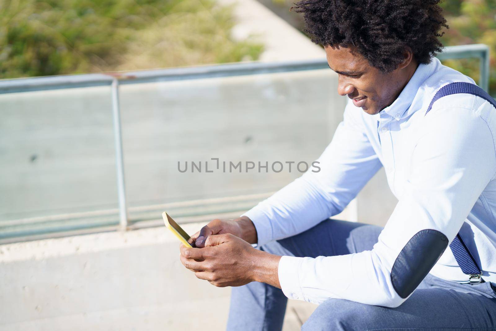 Black man with afro hairstyle using a smartphone sitting near an office building. by javiindy