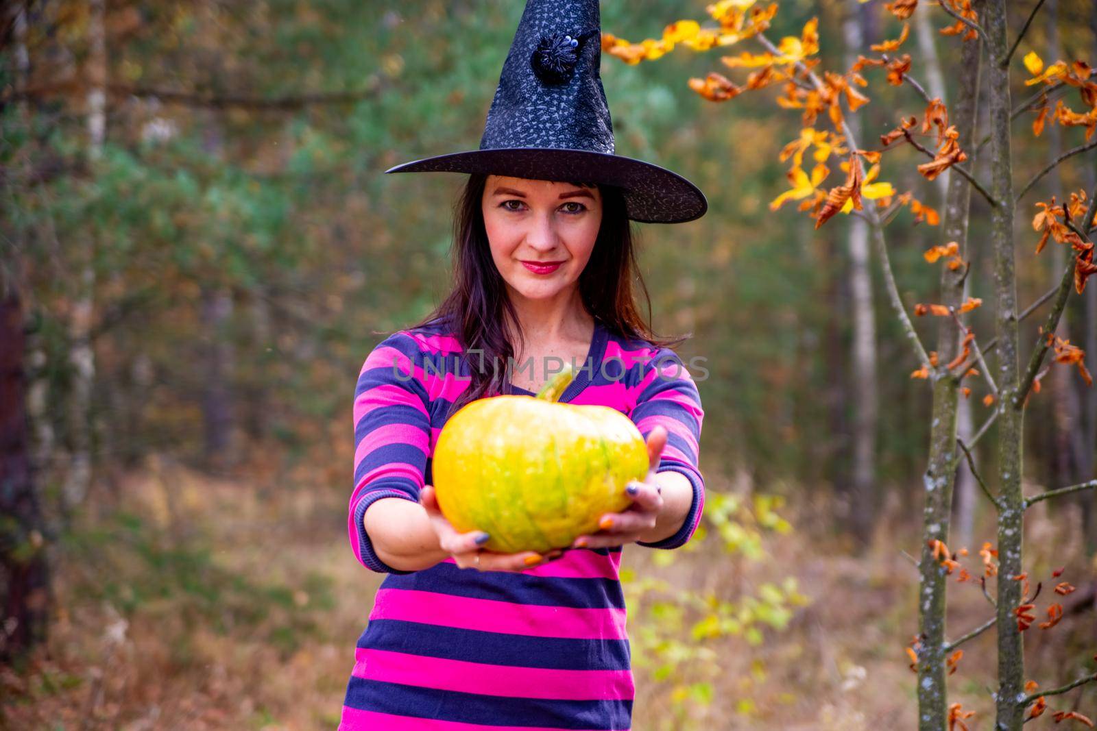 witch holds out a pumpkin to the camera in the autumn forest by Mariaprovector