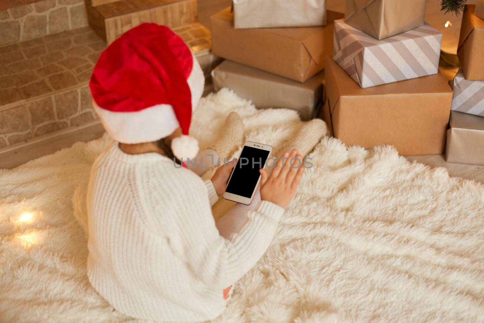 Little girl holding phone with blank screen while posing backwards, wearing white sweater and santa hat, sitting on floor on soft carpet. Merry Christmas and Happy Holidays! by sementsovalesia