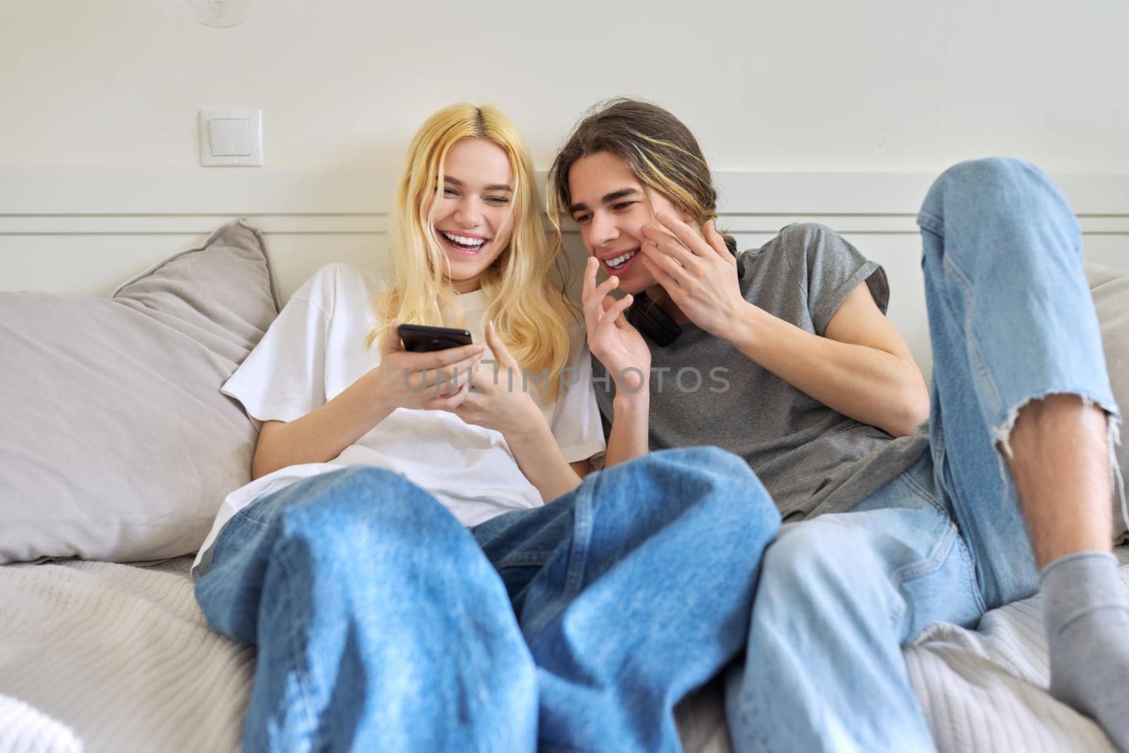 Couple of cheerful emotional teenagers friends having fun together, sitting on couch looking into smartphone. Adolescents male and female 15, 16 years old, technology, lifestyle, fun, friendship
