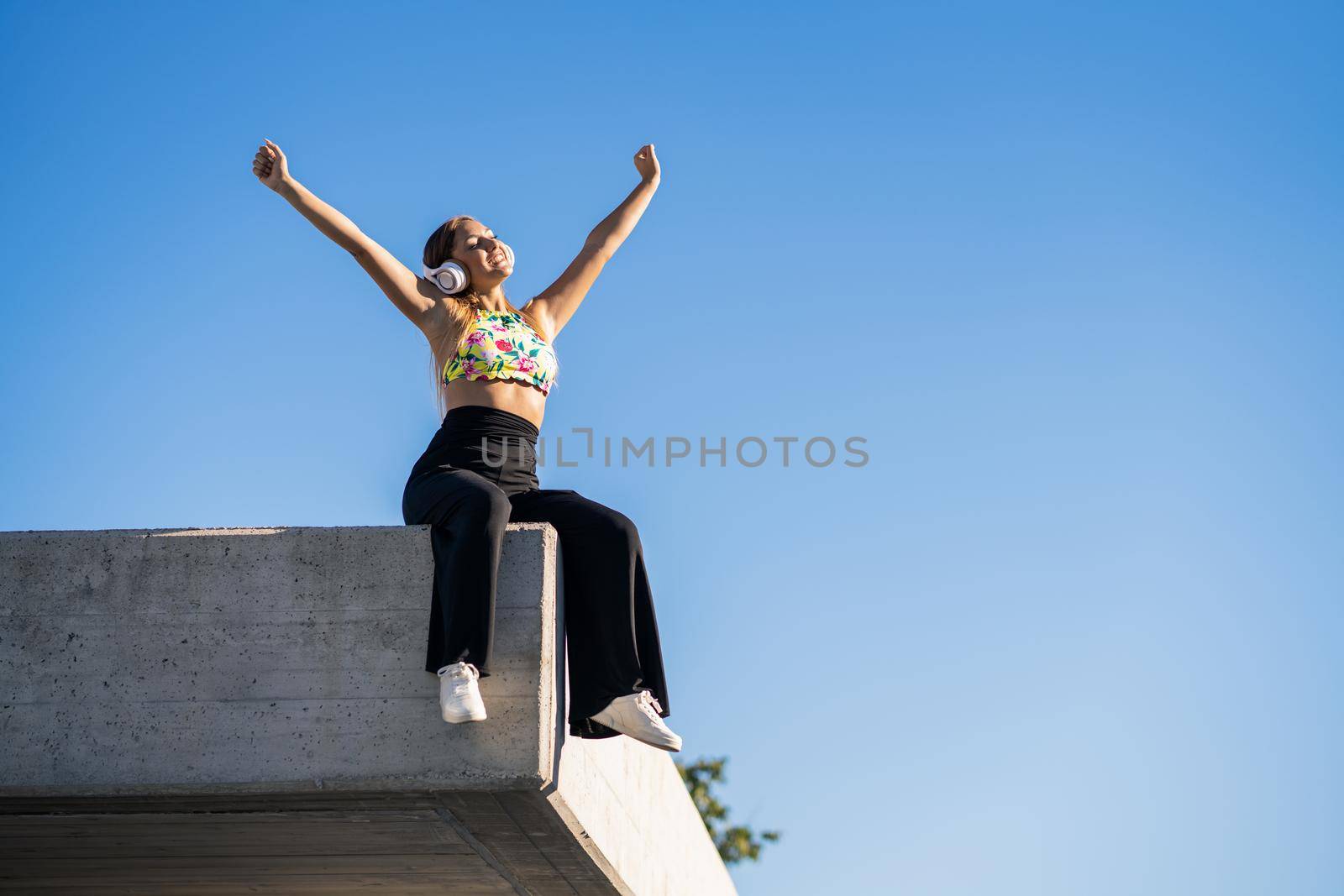 Young woman opening arms wearing headphones against blue sky.