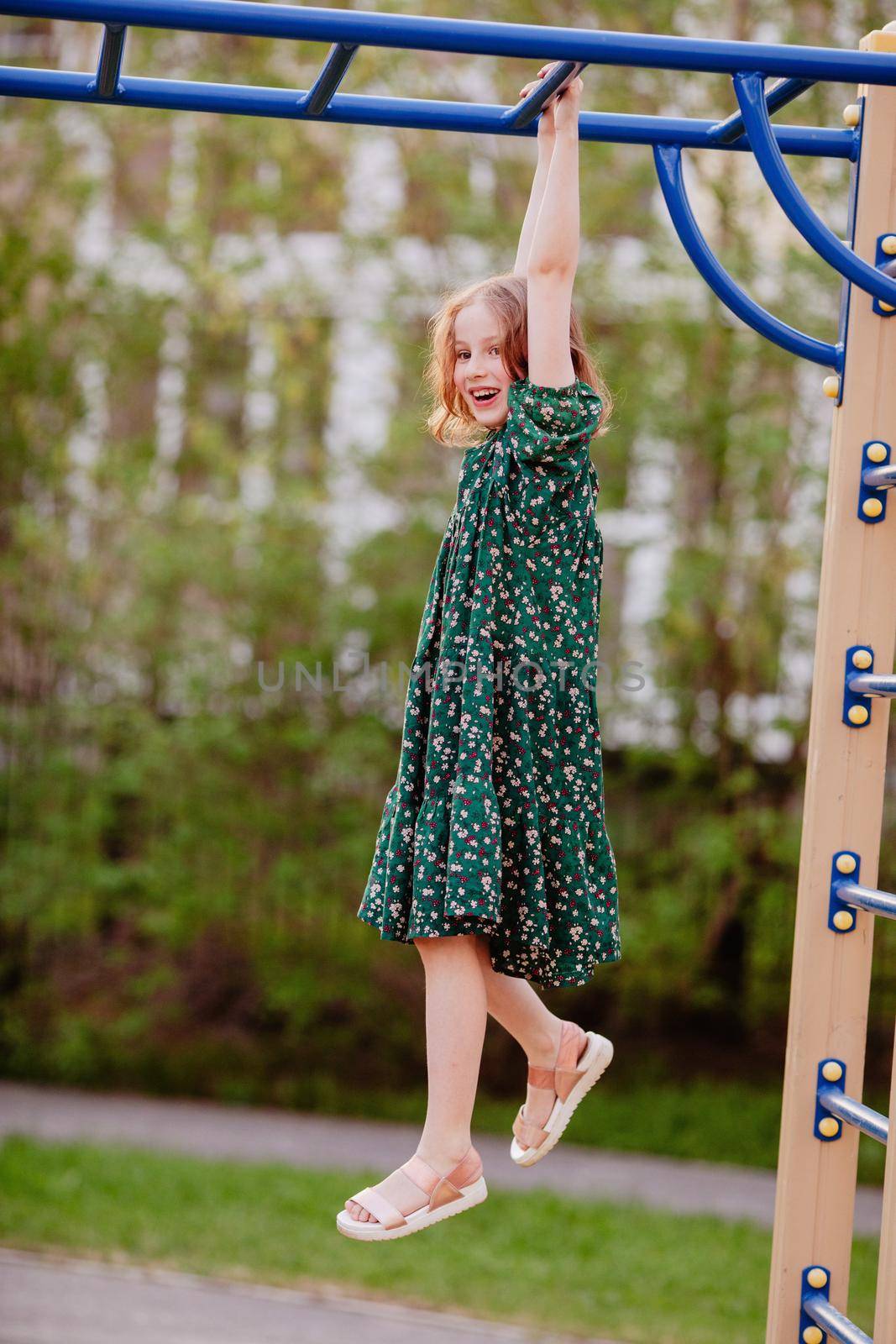Side view of carefree girl in dress hanging on metal monkey bars while having fun on playground in summer
