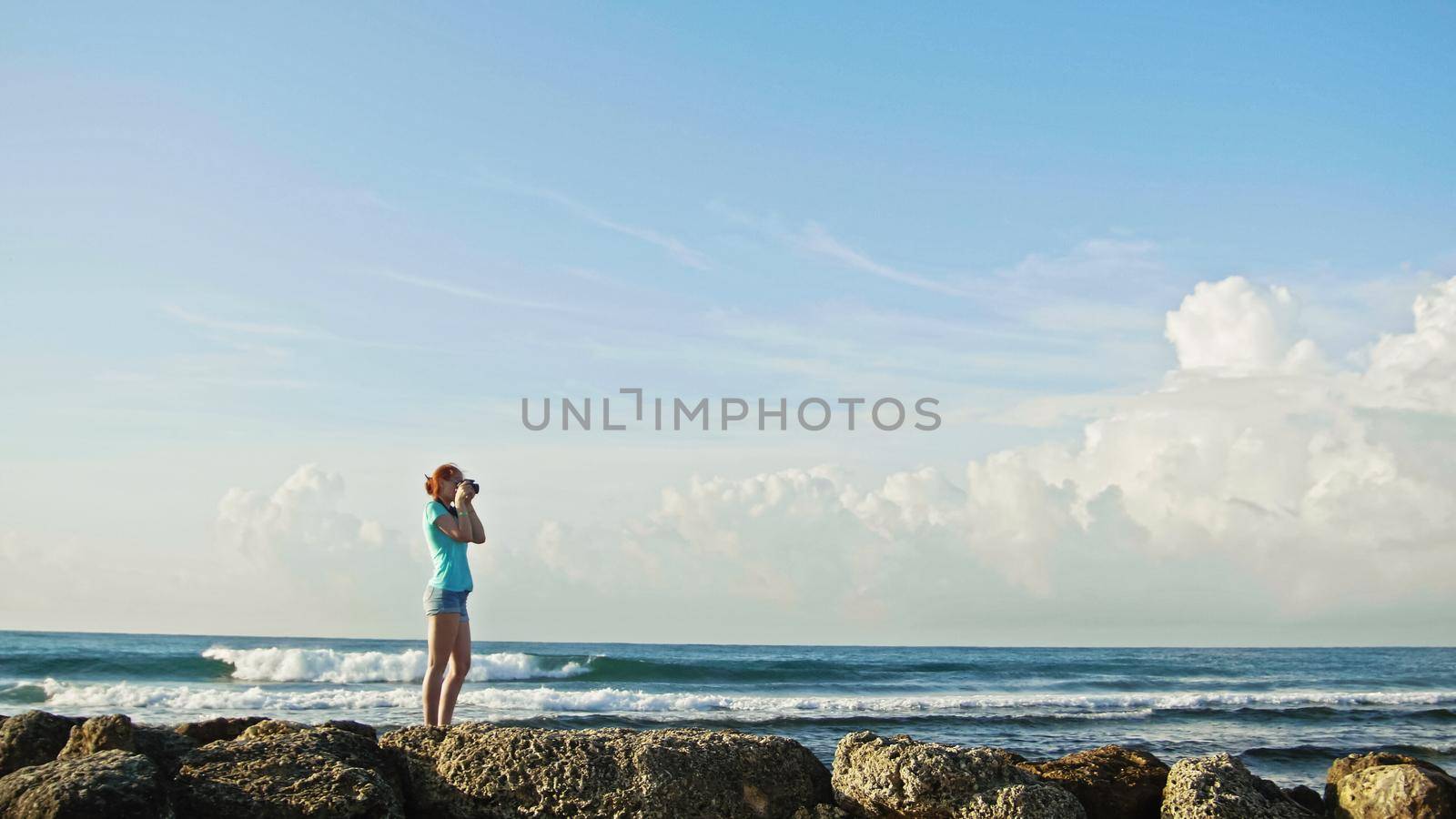 Young woman with red hair in glasses photographs sea standing on the rocks , beach of Dominican Republic, wide angle, Caribbean sea