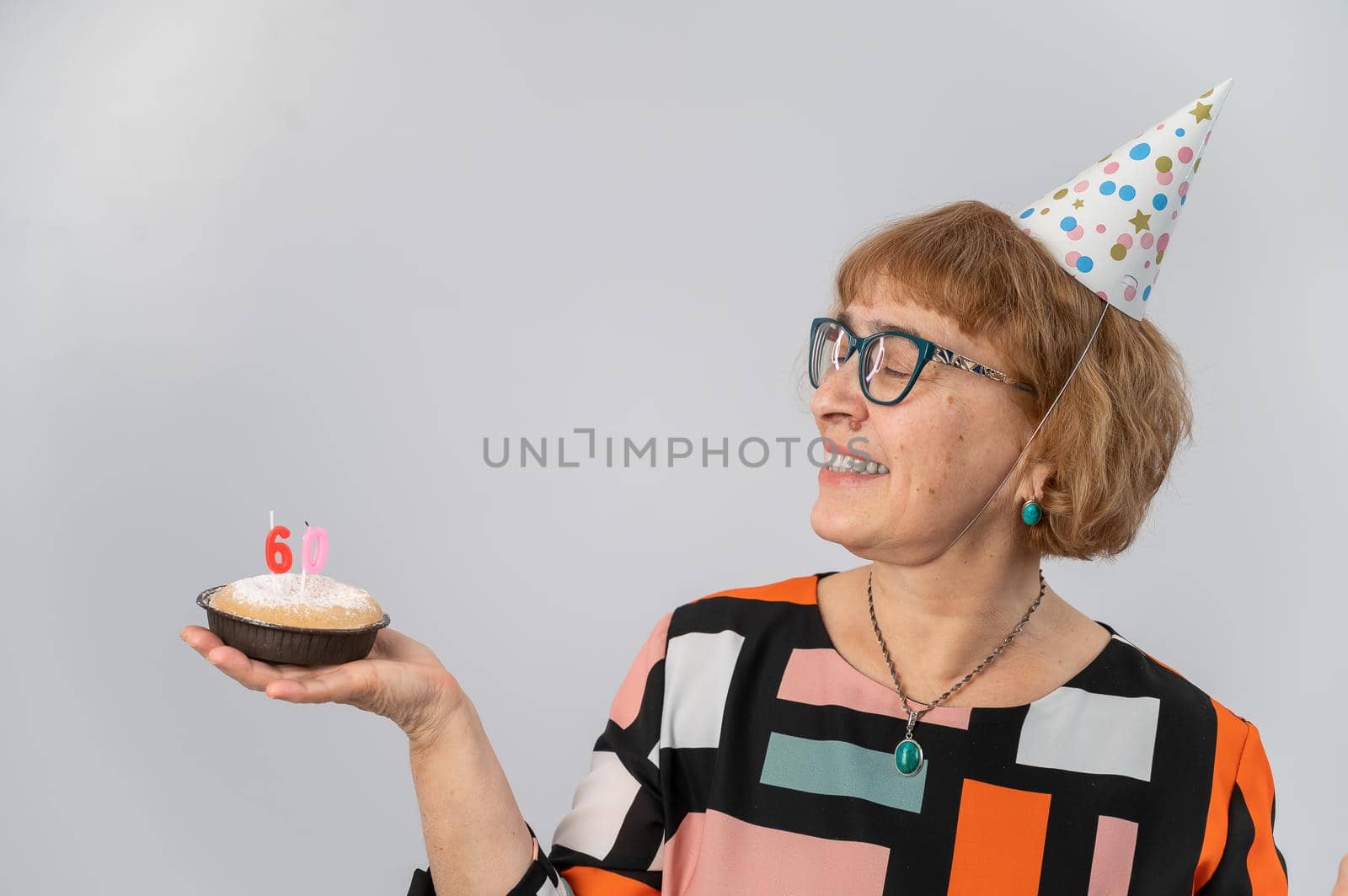 A portrait of a smiling elderly woman in a festive cap holding a cake with candles in the form of the number 60. Anniversary.