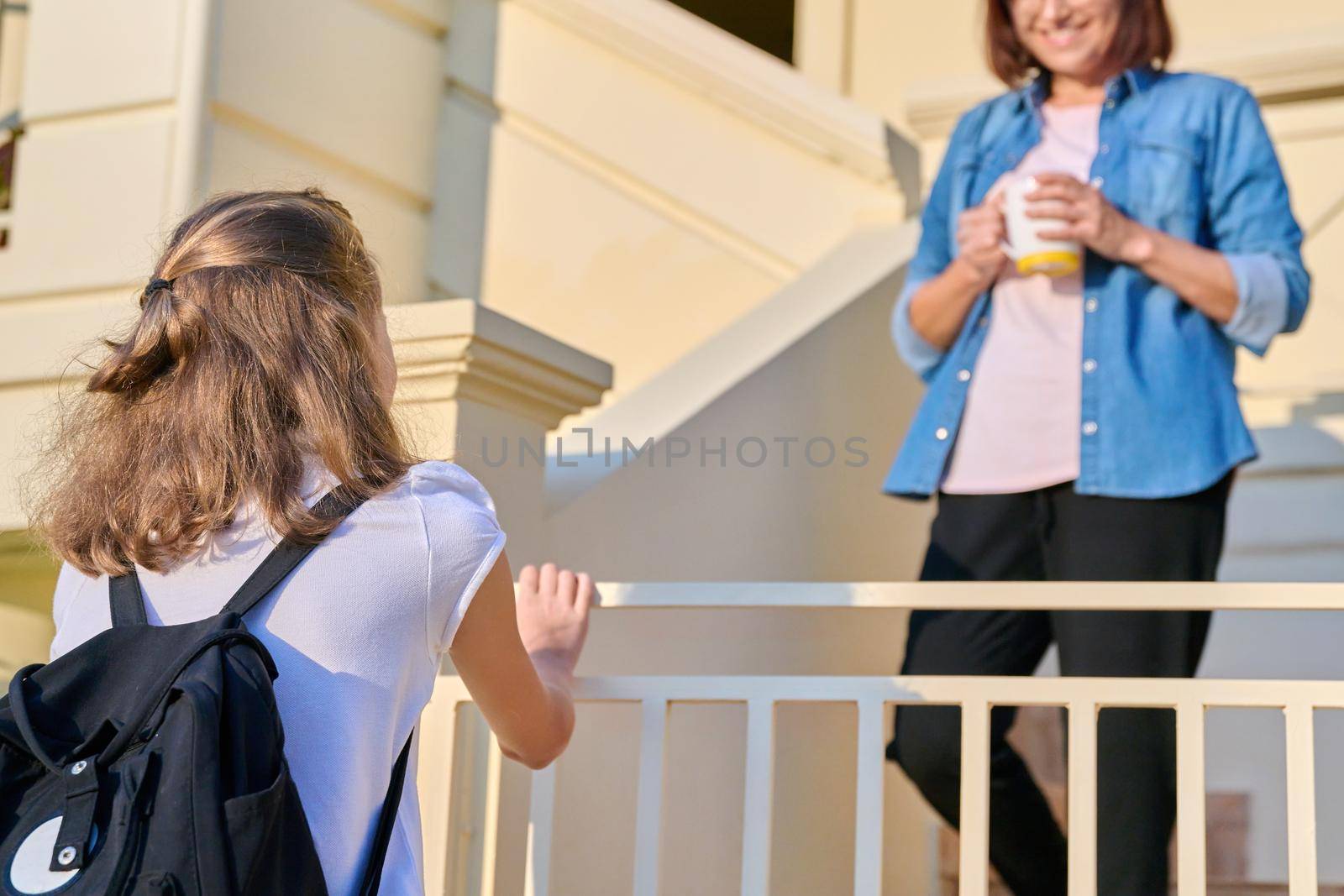 Girl student with backpack on porch of house with mother. Child going to school in morning, mom standing on outdoor steps of house saying goodbye and good day. Family, back to school, parent child