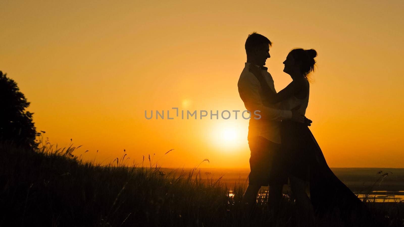 Young loving couple - young man and beautiful girl stands on high hill at sunset and have hugs and kiss, silhouette telephoto shot