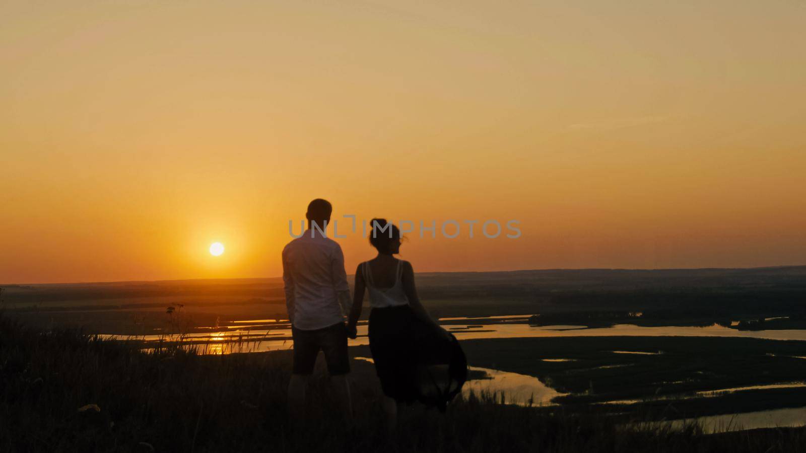 loving couple - brave young man and beautiful girl stands on high hill looking to sunset silhouette by Studia72